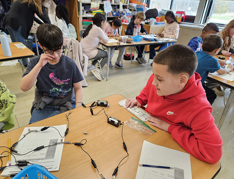 Two fourth-grade students sit at a desk that has lightbulb, and wires.