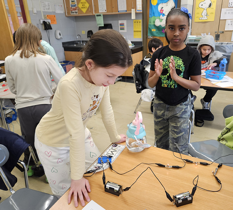 Two fourth-grade students stand at a desk that has lightbulb, and wires.