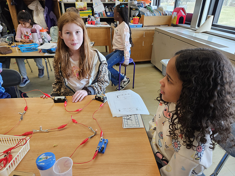Two fourth-grade students sit at a desk that has lightbulb, and wires.