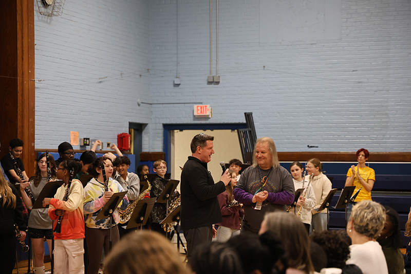 A man stands in front of a large group of fifth-grade kids talking. Behind him are kids with musical  instruments.