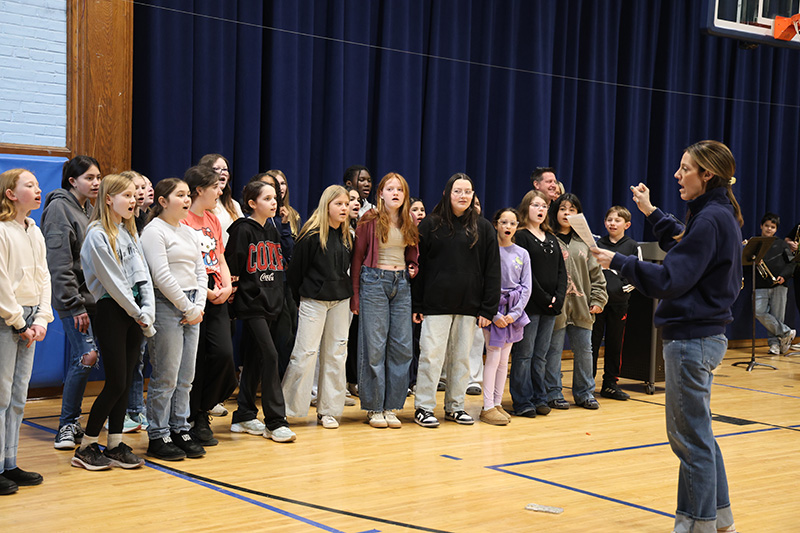 A group of middle school kids stand together and sing. There is a woman instructing them.