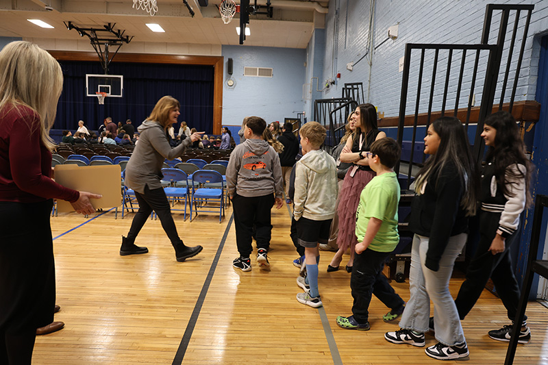 Fifth-grade kids stream into a gymnasium and a woman stops one to say hello.