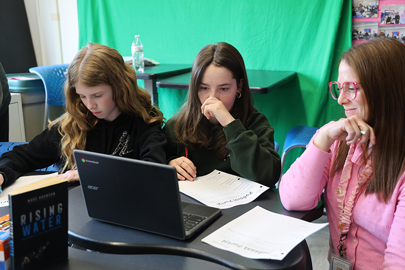 Two middle school girls work on their podcast projects while their teacher, a woman wearing pink, sits beside them.