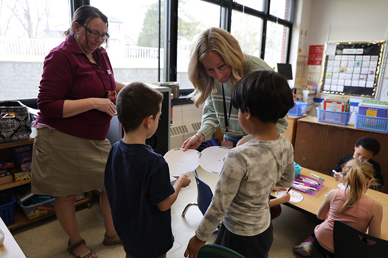 A woman looks on as another woman helps two children with their art projects.