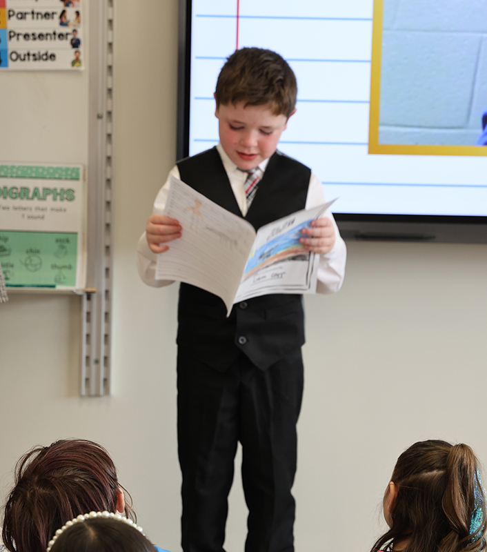 A young boy in a suit and tie reads from his story in front of his classmates.