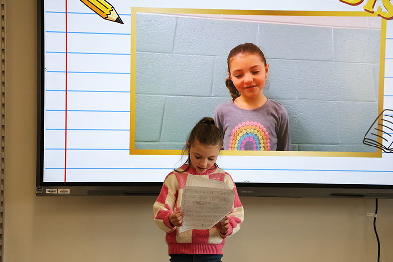 A young girl reads from her writing. Behind her is a video of her.