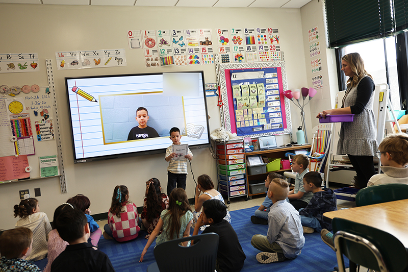 A young boy stands in front of a screen that has a picture of him on it. He reads a story he wrote from the paper n front of him. Many of his classmates are sitting in front of him listening.