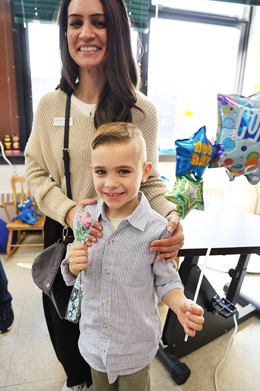 A first-grade boy smiles as he stands with a woman, who has long dark hair. She has her arms around him.