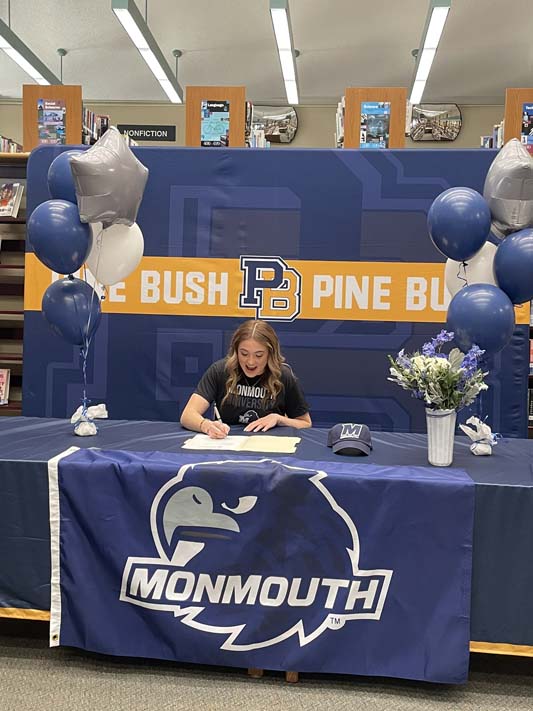 A young woman with long blonde hair sits at a table that says Monmouth and signs a paper. There are balloons on the table too. 