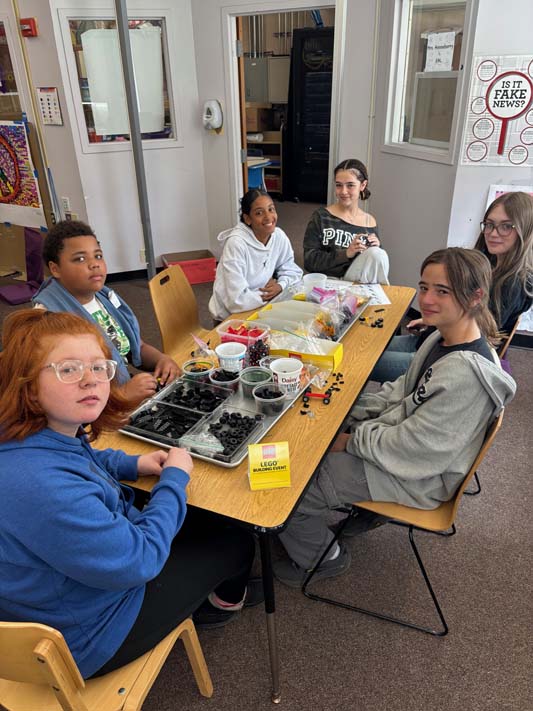 A group of six middle school kids sit around a table making things with Legos.