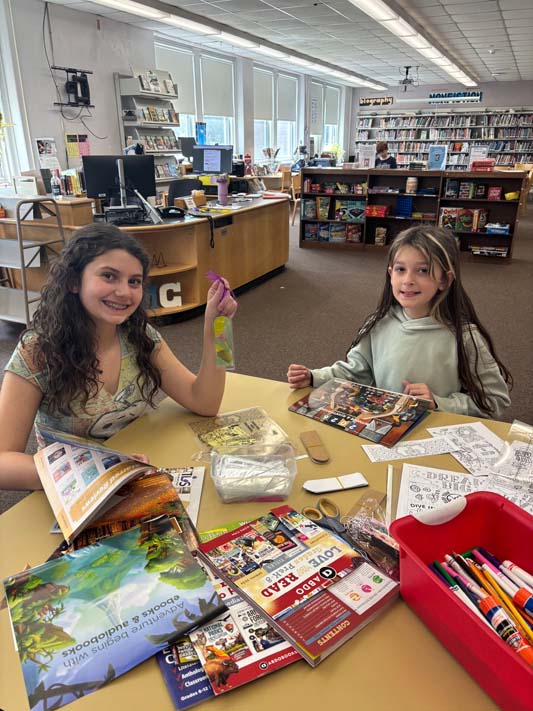 Two middle school girls smile as they use markers and craft items to make a book mark.