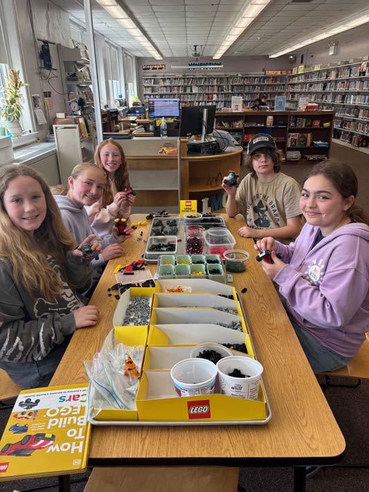 A group of five middle school kids sit around a table making things with Legos.