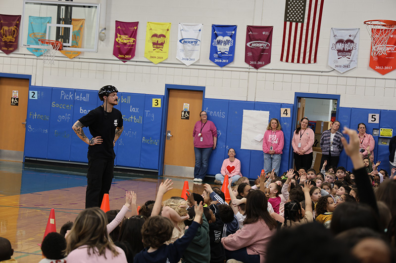 A man in black athletic wear and a bicycle helmet stands in a gymnasium and talks to kids, some of whom are raising their hands.