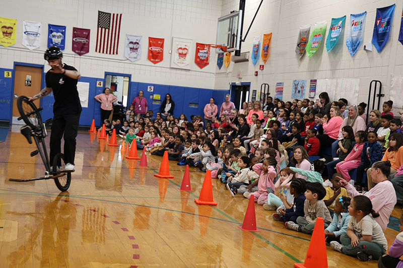 A large group of elementary students sit on bleachers and watch as a man rides his bike and talks to them.