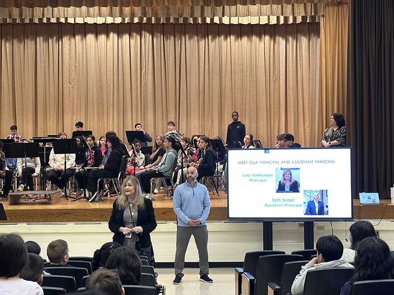 A man and woman stand in front of a large group of students with a screen to the right.