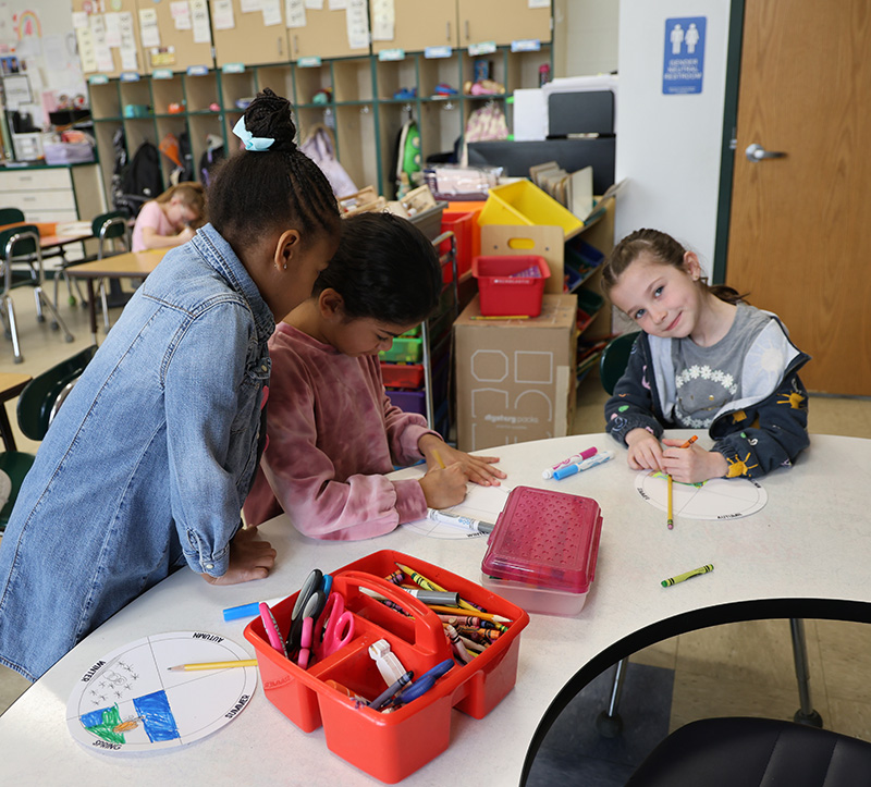 Three first graders sit at a table drawing art projects.