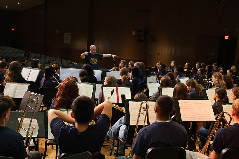 A man stands with his arms out conducting a large group of high school and middle school musicians on a stage.