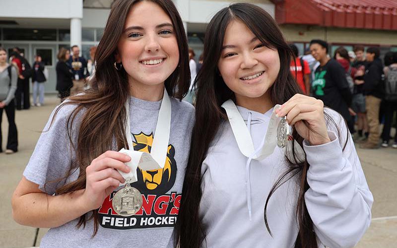 Two young women, both with long dark hair, smile and hold up the silver medals they won.