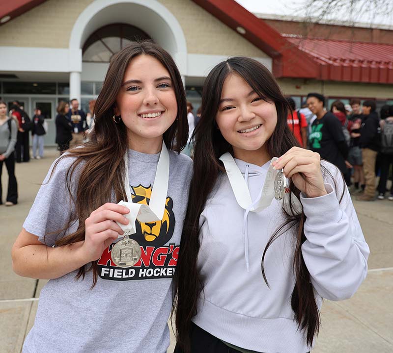 Two young women, both with long dark hair, smile and hold up the silver medals they won.