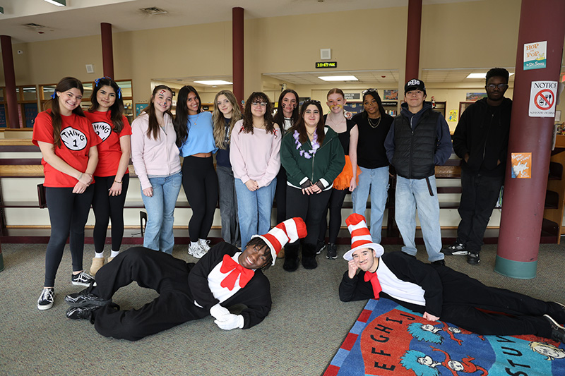 A group of  14 high school kids. Two are dressed as the cat in the hat and are laying across the floor in front.