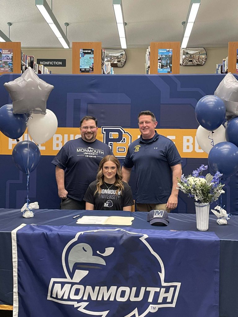 A high school girl sits at a table with a blue tablecloth that says Monmouth on it. Two men are standing behind her. Everyone is smiling.