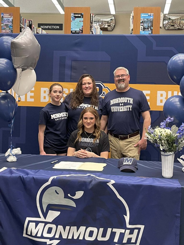 A high school girl sits at a table with a blue tablecloth that says Monmouth on it. A man, woman and girl are standing behind her. Everyone is smiling.