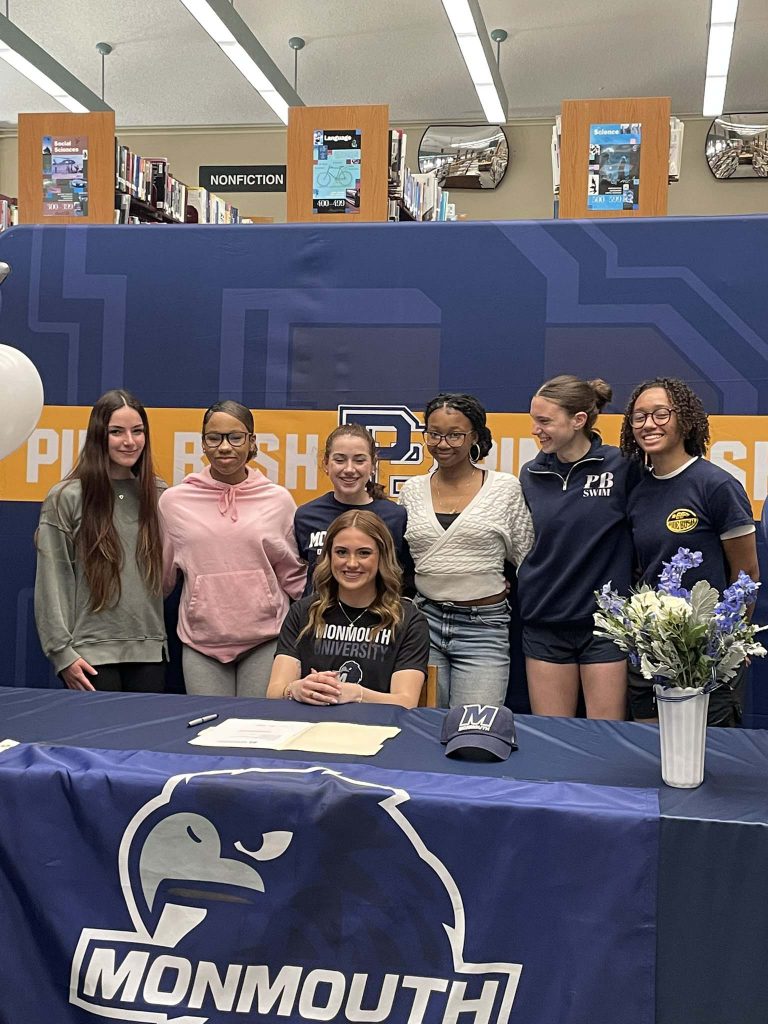 A high school girl sits at a table with a blue tablecloth that says Monmouth on it. TA group of high school girls are standing behind her. Everyone is smiling.
