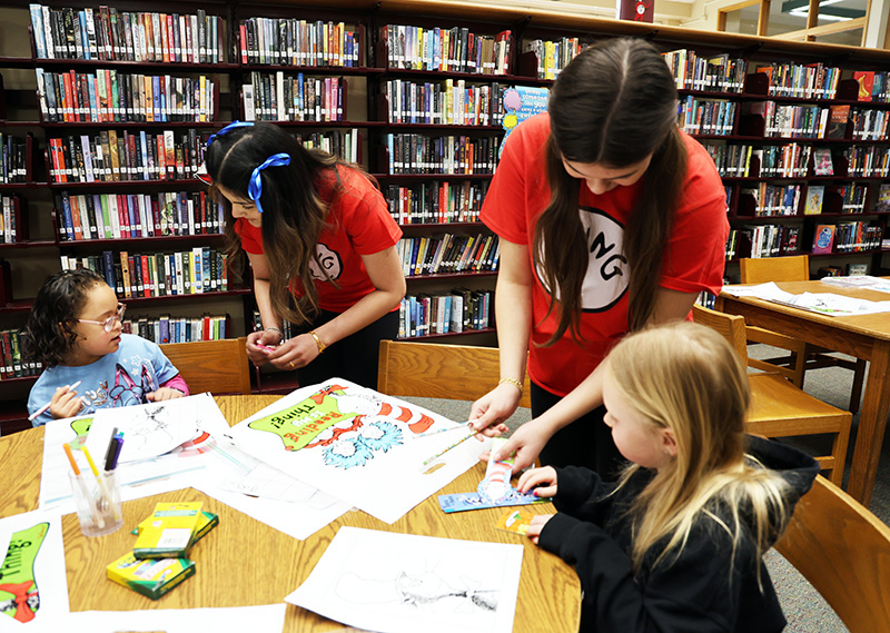 Two high school girls wearing red tshirts that say Thing 1 and Thing 2 help kindergarten kids color.