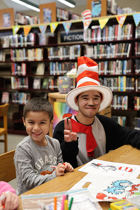 A high school kid dressed as the cat in the hat gives a peace sign. He is sitting with a kindergarten kid who is smiling.