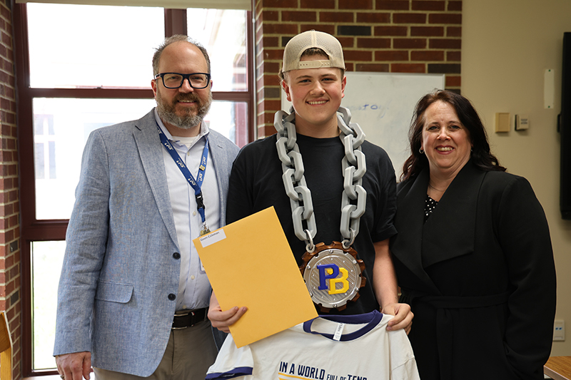 A young man stands between a man and a woman. they are all smiling. He is wearing a very chunky chain with a huge PB medallion on it.
