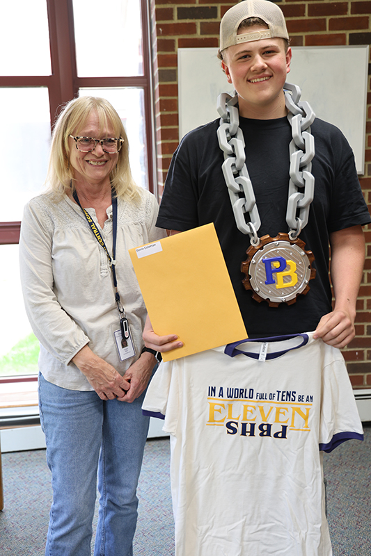 A woman with chin-length blonde hair and glasses stands with a high school kid who is holding a blue and gold tshirt. He is also wearing a huge chunky chain with a PB emblem on it.