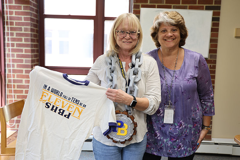A woman with chin-length blonde hair holds a blue and gold tshirt that says "In a world full of tens be an Eleven PBHS." She is standing with another woman. Both are smiling. 