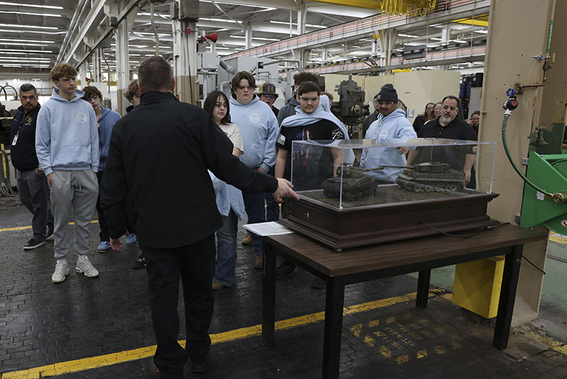 A group of high school students listen as a man talks to them in a factory.