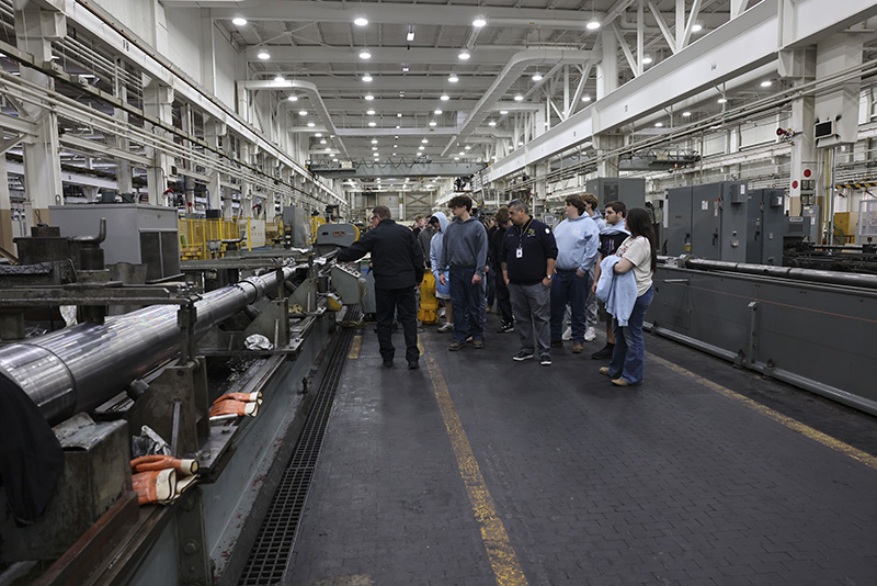 A group of high school students listen as a man talks to them in a factory.