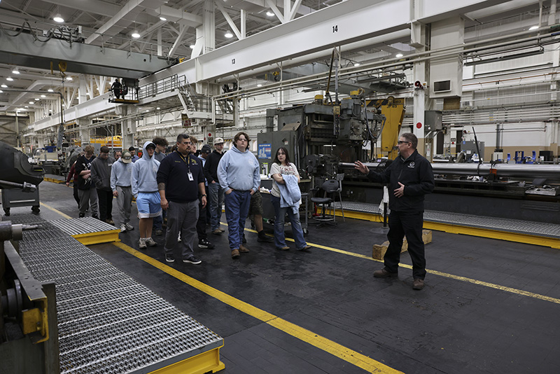 A group of high school students listen as a man talks to them in a factory.