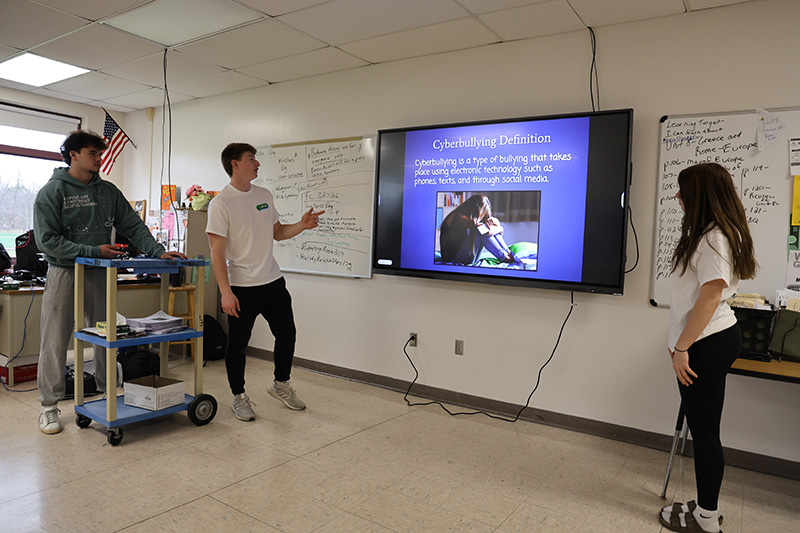 A monitor in the center. Three high school kids are using it to give a presentation to kids in a classroom.