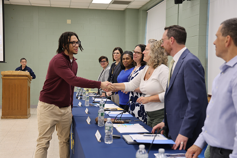 A young man wearing a maroon shirt and khaki pants shakes hands with a woman as other adults wait to shakes hands as well.