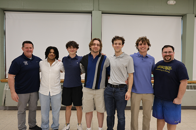 A group of five high school boys stand together smiling, A man stands on the right of them and another on the left.