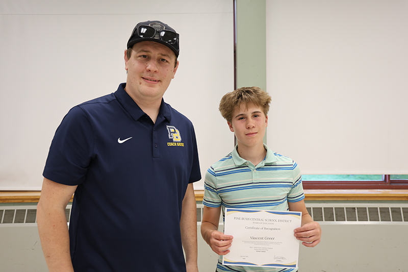 A man wearing a Pine Bush wrestling shirt smiles and stands with a young man who holds a certificate. 