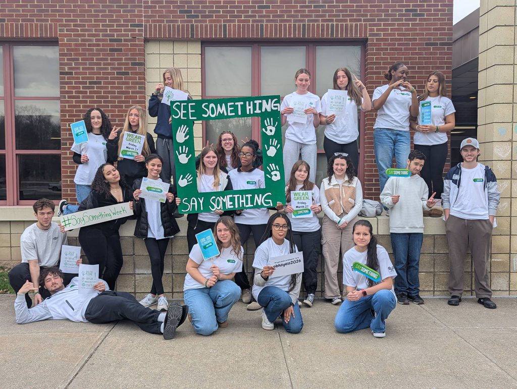 A group of 20 high school students stand around with their green and white Sandy Hook Promise shirts on. Some are holding a square photo booth that says See Something Say Something.