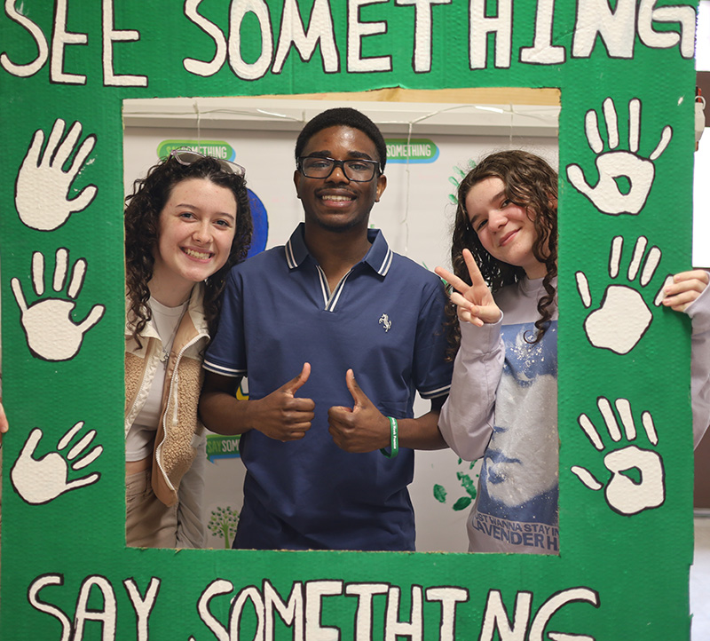 Three high school kids smile as they hold up a photo booth sign that says See Something Say Something.