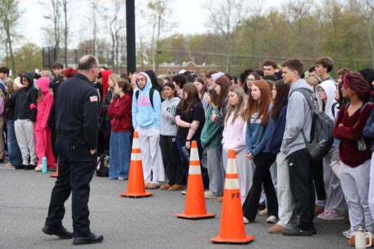 A large group of high school students stand around listening as a police officer talks.