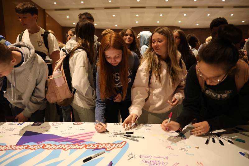 A group of high school kids lean over a large poster to sign a pledge.
