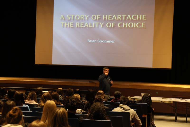 A man stands in an auditorium in front of many high school kids. Behind him is a screen that says A story of heartache The reality of choice