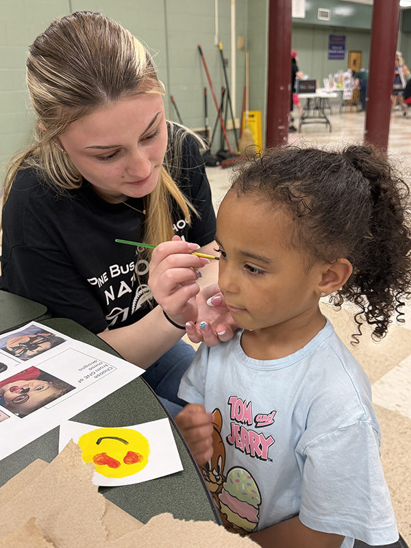 A young girl sits as a high school girl paints her face.
