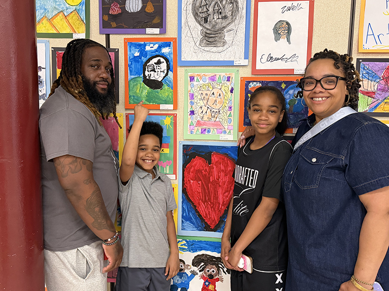 A family of two adults and two kids stand and smile at a wall of art. The youngest kid points to his artwork.