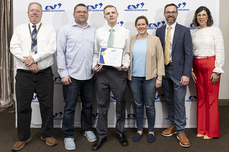 Six people stand together smiling. They are in front of a background that says OU BOCES. The high school kid in the center holds a certificate.