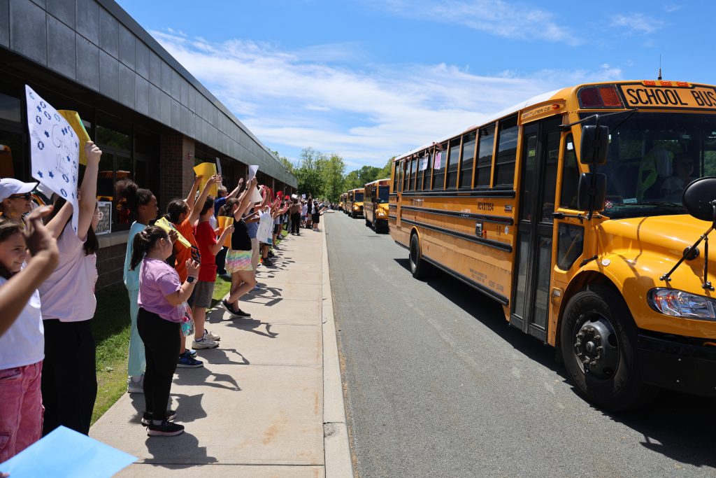 A line of elementary students cheer and watch as yellow school buses drive by.