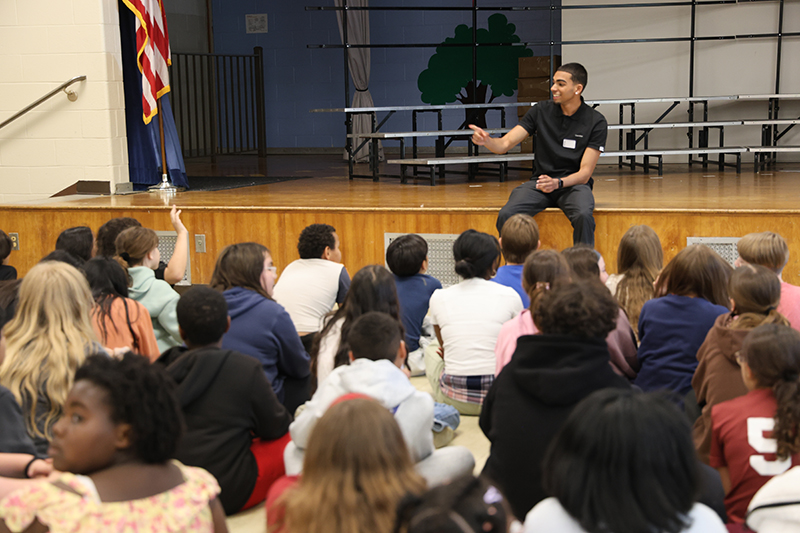 A young man sits on th eedge of a stage and talks to a group of fifth-grade kids sitting on the floor in front of him.