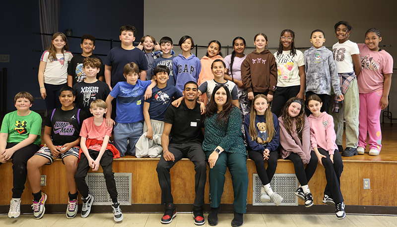 A group of about 25 fifth-grade students stand and kneel around a young man and a woman who are sitting on the edige of a stage.